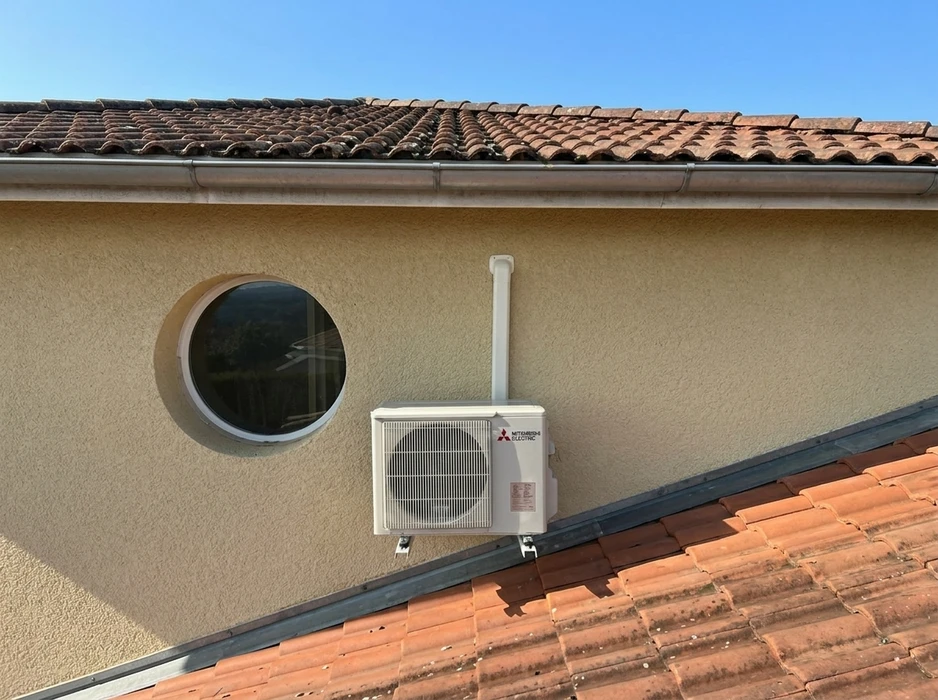 Unité extérieure de climatisation Mitsubishi Electric installée en hauteur sur un mur de façade enduite, posée sur supports métalliques sur toiture tuiles canal
